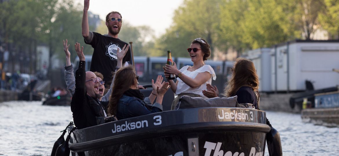 Three men and three women popping champagne on boat on amsterdam canals